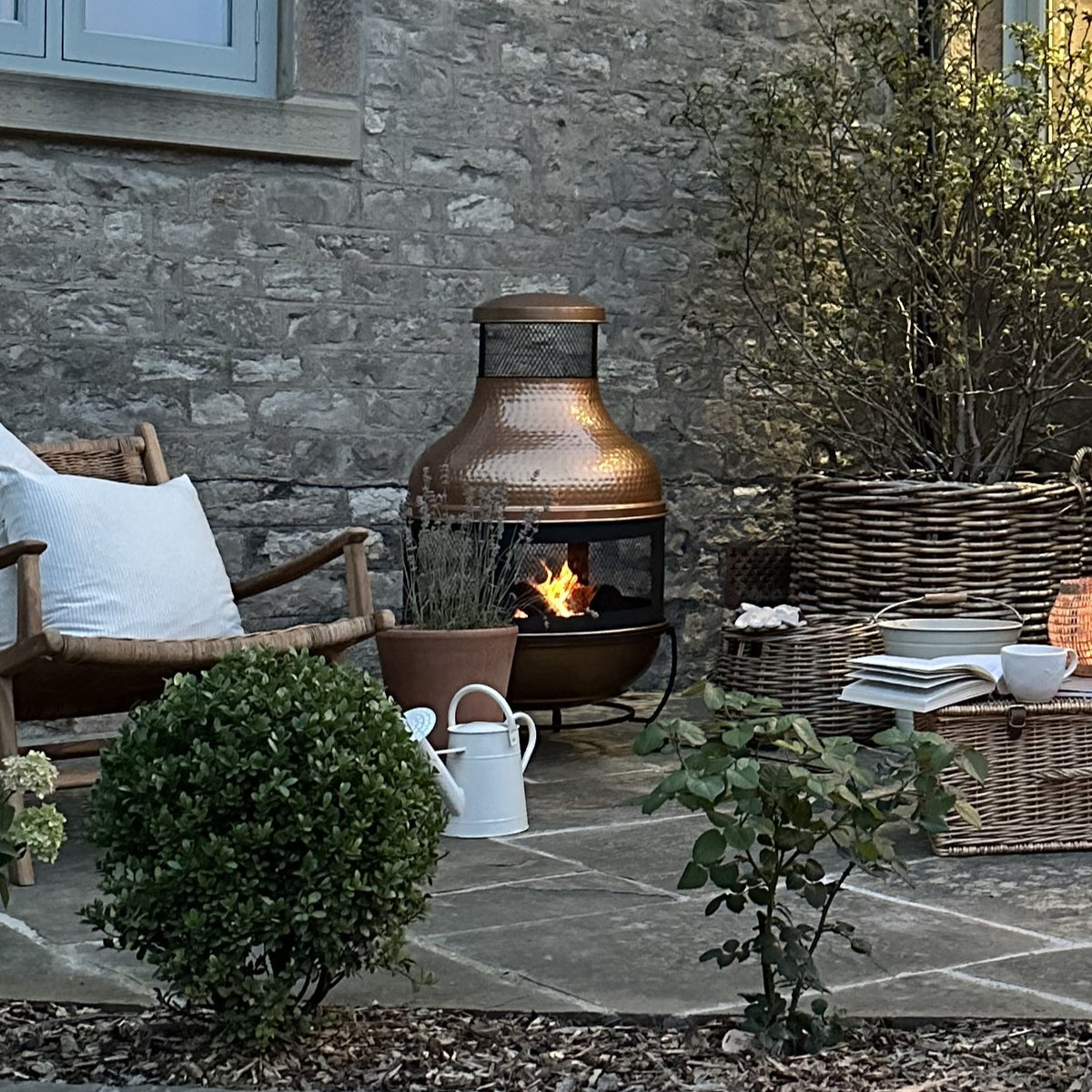 Large copper fire pit lit, outside on flagstone floor next to rattan chair, behind bushes