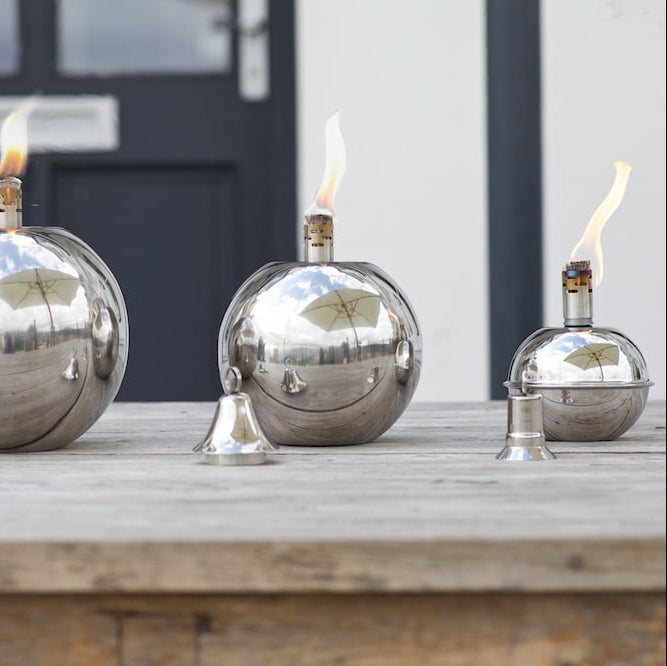 Three polished silver oil lamps with live flames, arranged on a weathered wooden surface outdoors in front of a dark door.