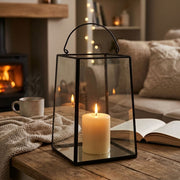 A black metal lantern with clear glass panels and a curved top handle sits on a wooden table in a cozy living room. A lit cream pillar candle is placed inside the lantern, with a mug, blanket, lit fireplace.