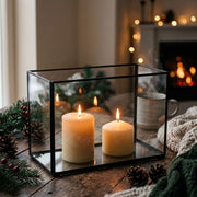 A black metal and glass rectangular lantern with two lit cream pillar candles inside, placed on a rustic wooden table with pine branches, pinecones, and a cozy fireplace in the background.