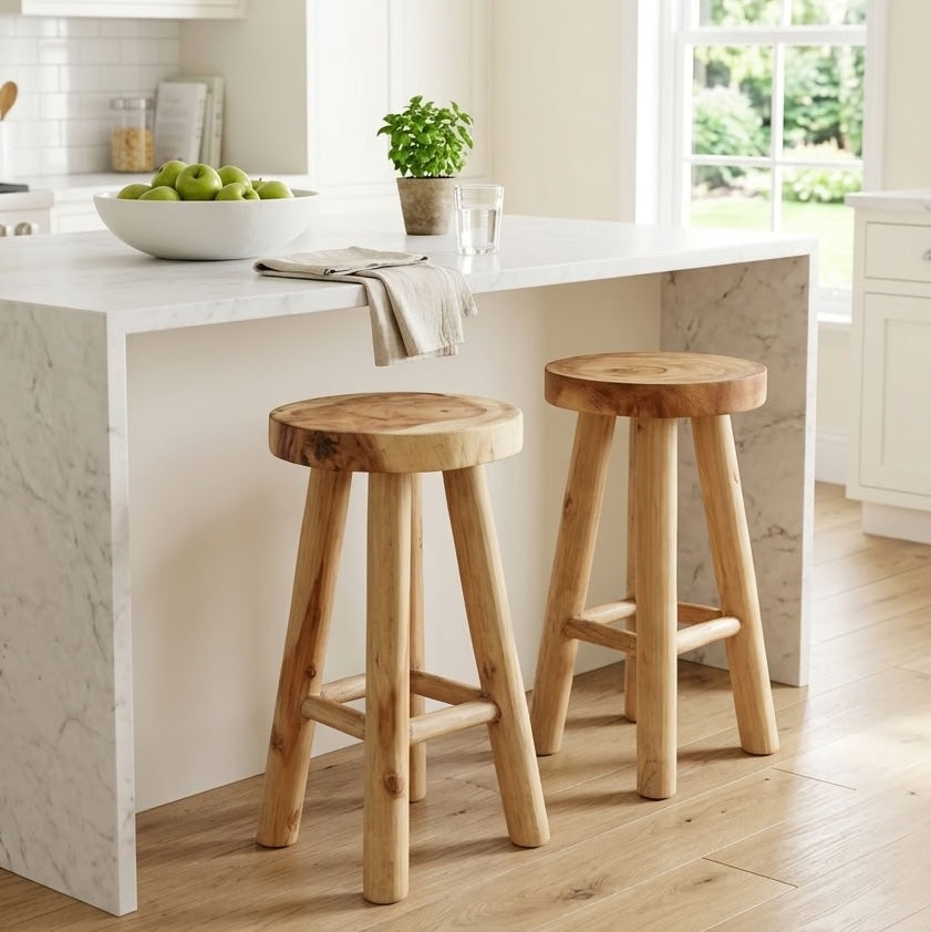 Two rustic wooden bar stools with thick round seats and four legs, positioned in front of a white marble kitchen island in a bright, modern kitchen.