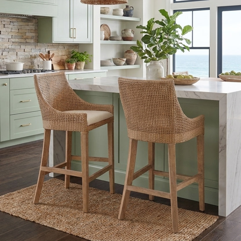 Two woven rattan bar stools with mango wood legs and cream cushions, positioned at a kitchen island with pale green cabinets and a white marble countertop.