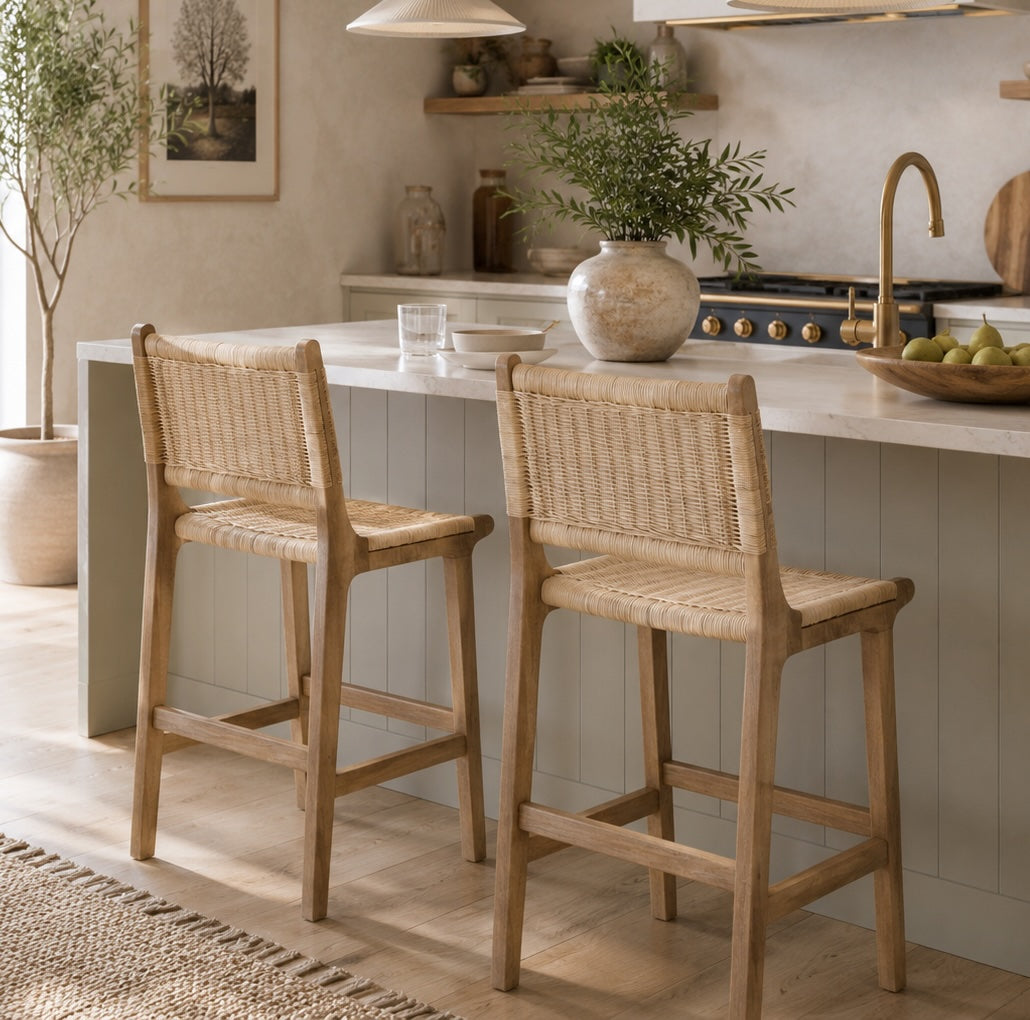 Two wooden and rattan bar stools with woven rattan seats and backs, positioned at a kitchen island in a bright, modern kitchen setting. Wicker rug and plants.