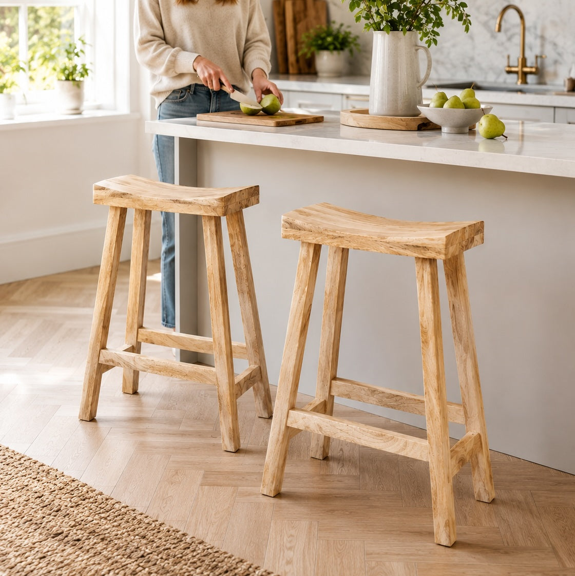 Two natural wood bar stools in a bright kitchen setting, with a person partially visible behind a counter cutting fruit.