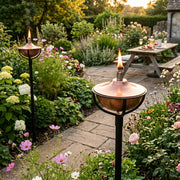 Two lit copper garden torches with black metal stands are placed on a stone pathway in a lush, flower-filled backyard garden. A wooden picnic table is visible in the background.