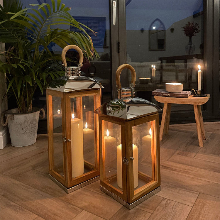 Two wooden lantern candles with glass panels and warm glowing flames, styled on a wooden floor in a modern interior setting with a potted plant and glass doors.