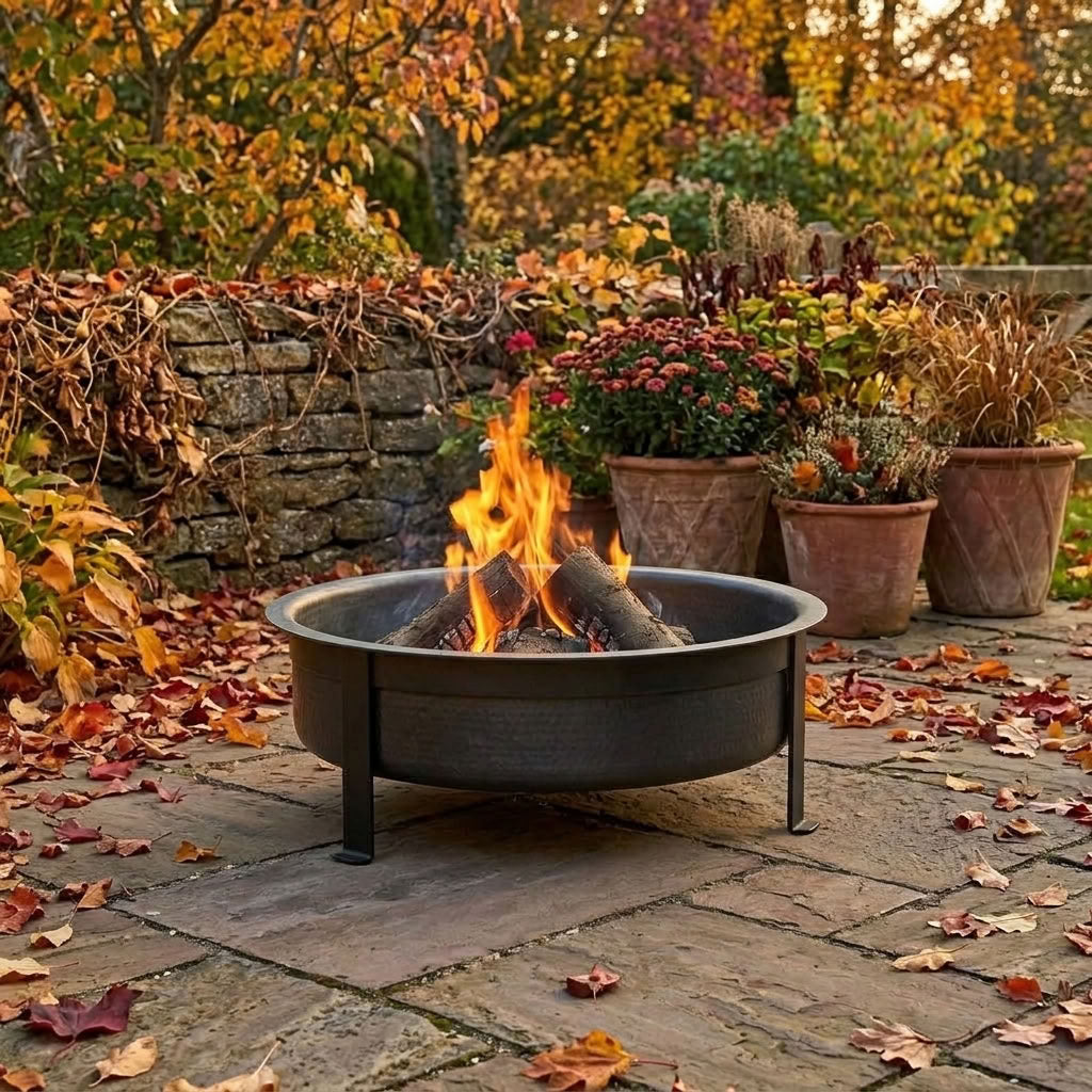 A round black metal fire pit with a fire burning inside, set on a stone patio in an outdoor garden with autumn foliage and potted plants.