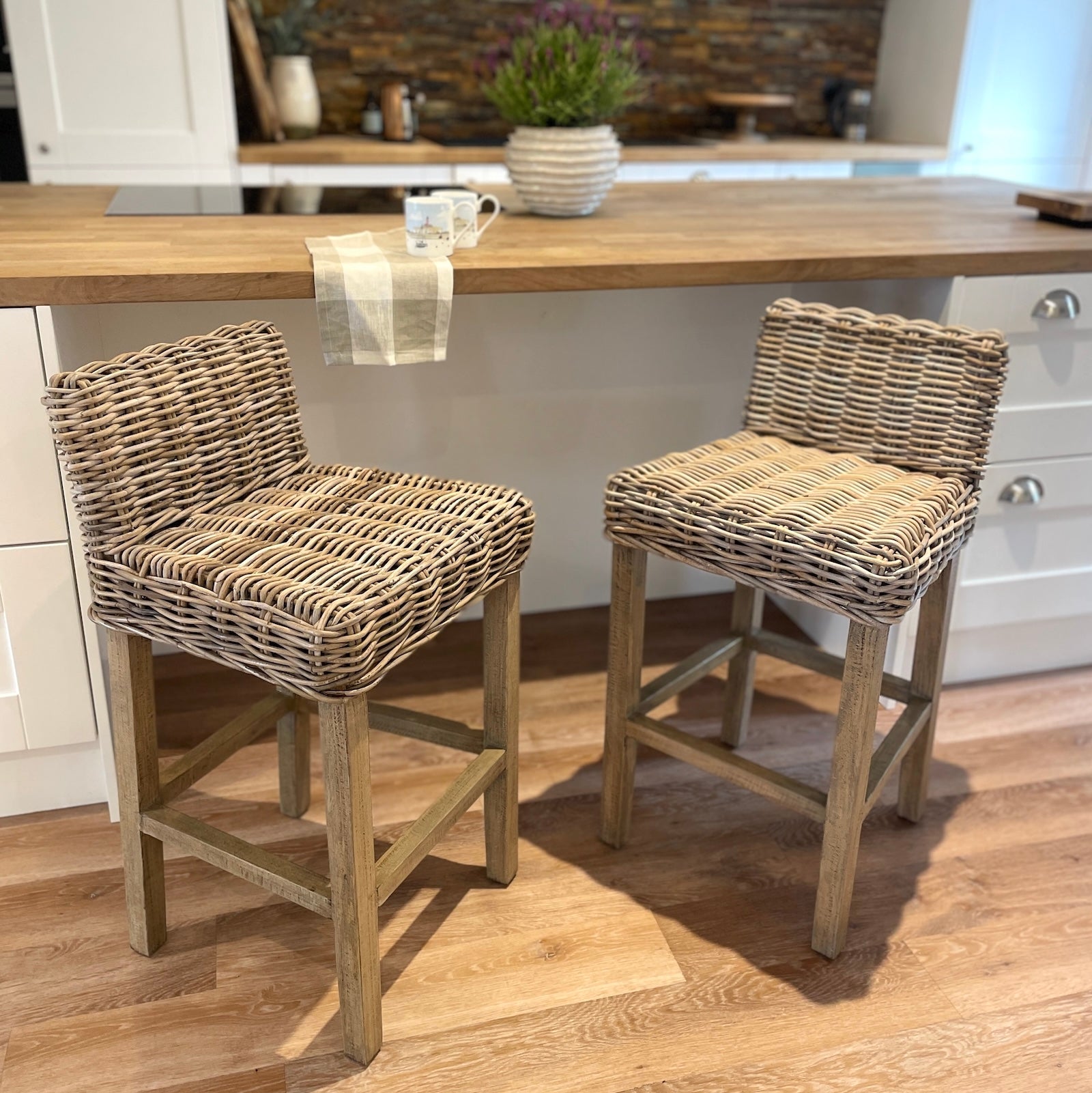 Two woven wicker bar stools with light wood legs, positioned in a kitchen setting in front of a wooden island counter.