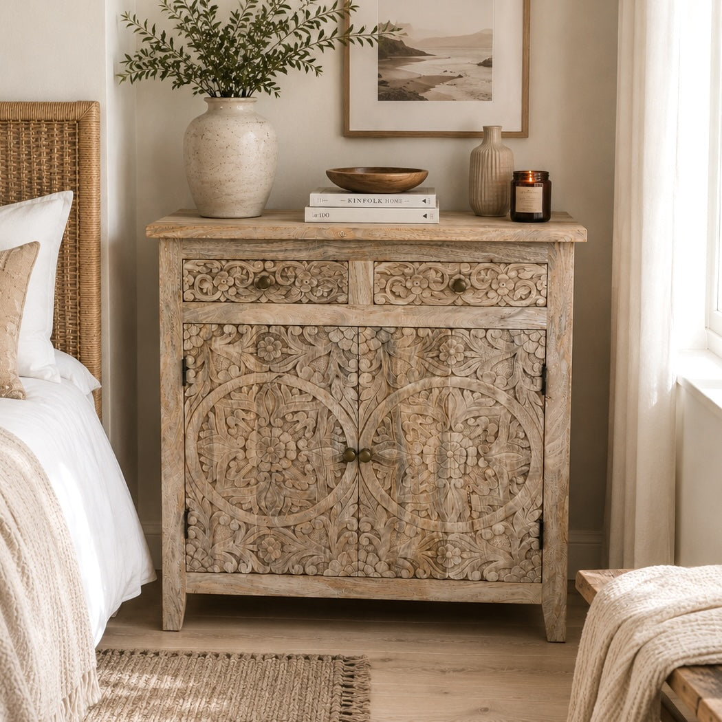 A carved wooden sideboard with two drawers and two doors, styled in a warm bedroom setting next to a bed with white linens.