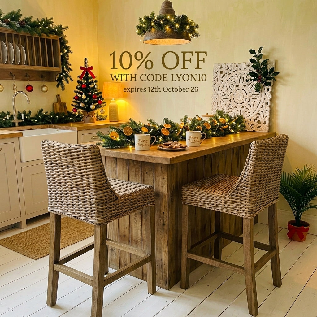 A rustic wooden bar-height island table with two woven wicker bar stools in a decorated holiday kitchen. The scene includes Christmas garlands, a small decorated tree, mugs on the table, and promotional text reading '10% OFF WITH CODE LYON10 expires 12th October 26'.