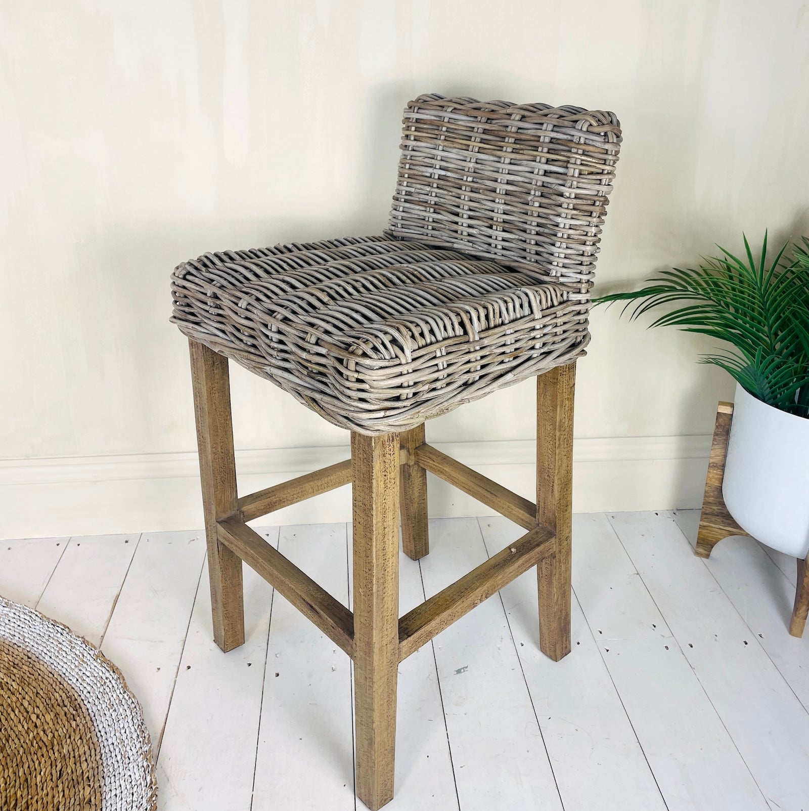 A rustic wicker barstool with a wooden base, set against a cream wall and white floor, with a potted plant in the background.