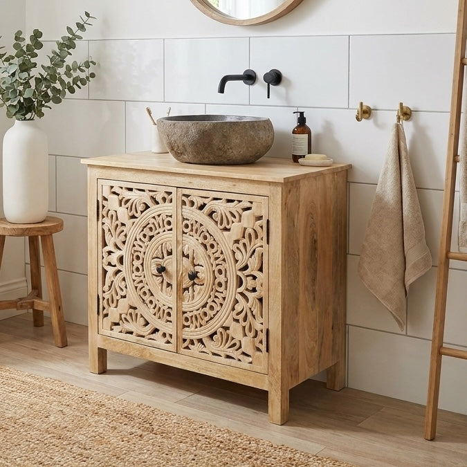 A light natural wood bathroom vanity cabinet with intricate carved doors, styled with a stone vessel sink, black faucet, and a beige towel in a bright bathroom setting.