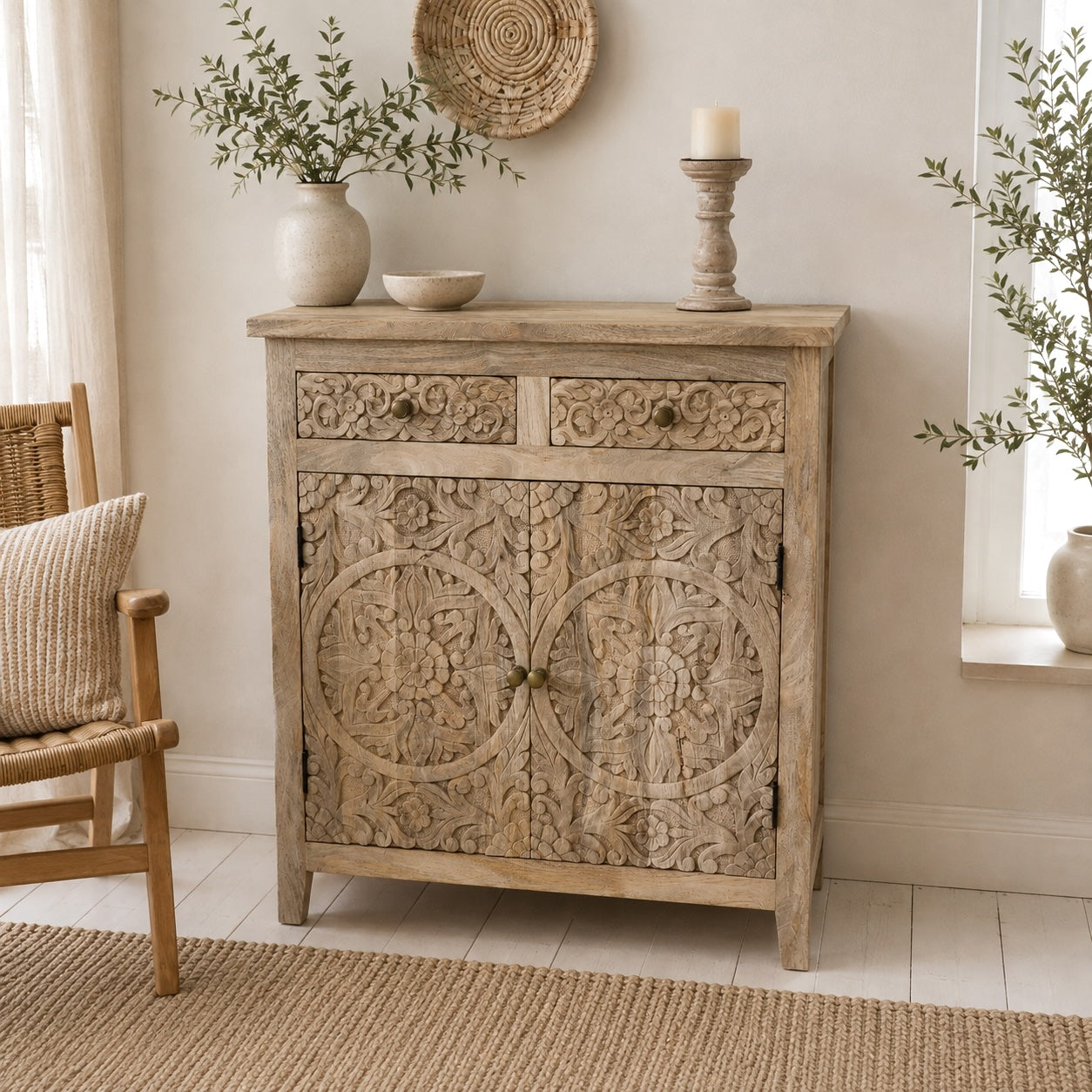 A carved wooden sideboard cabinet in a rustic light beige finish, styled in a bright room with a woven chair, potted plants, and decorative items on top.