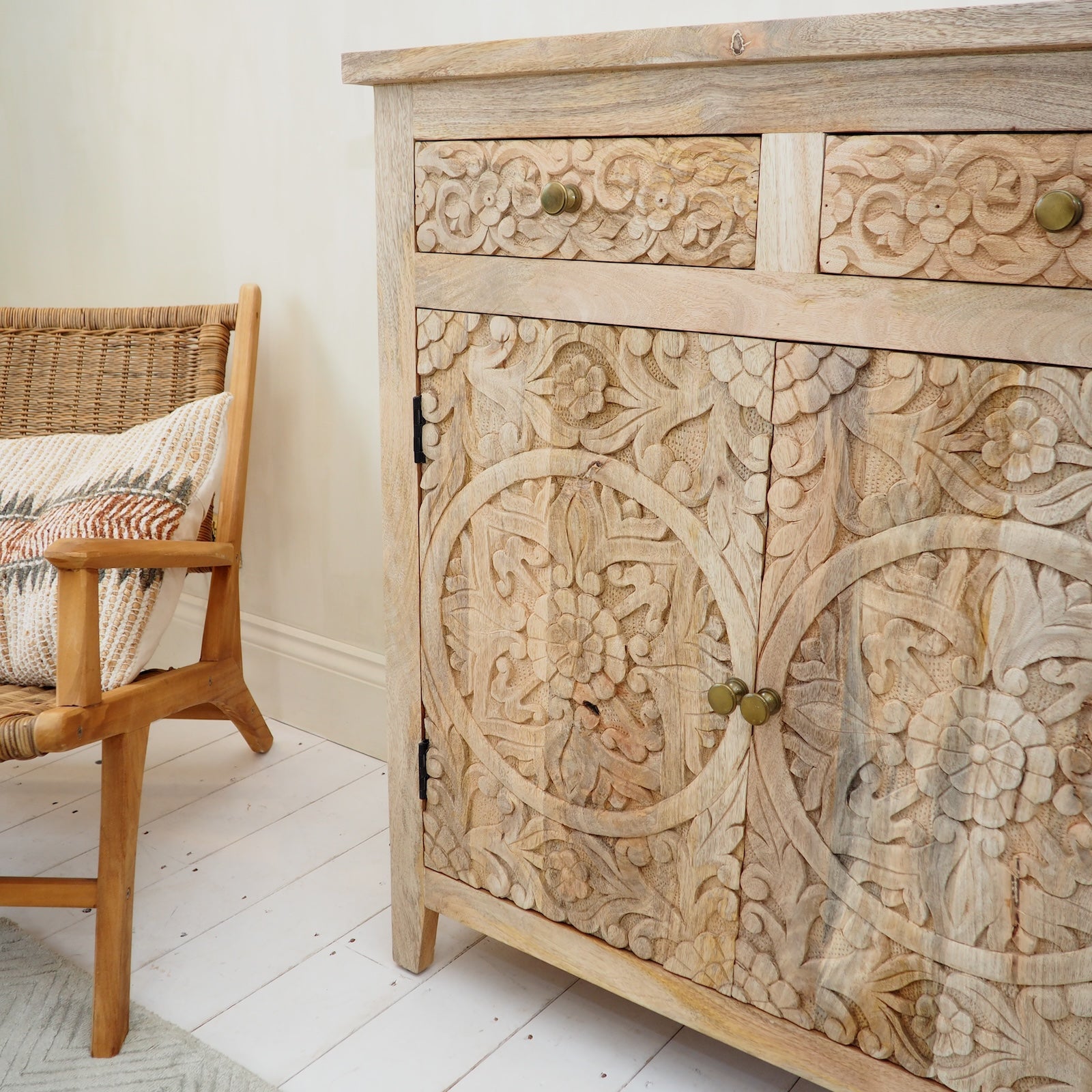 A carved wooden sideboard cabinet with floral detailing and brass knobs, shown in a styled room next to a woven chair.