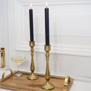 Two black taper candles in ornate gold candlesticks, displayed on a wooden serving tray with a coupe glass against a white paneled wall.