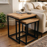A pair of sold wood square nesting side tables with wood tops and black metal frames, styled in a cosy living room next to a cream sofa.