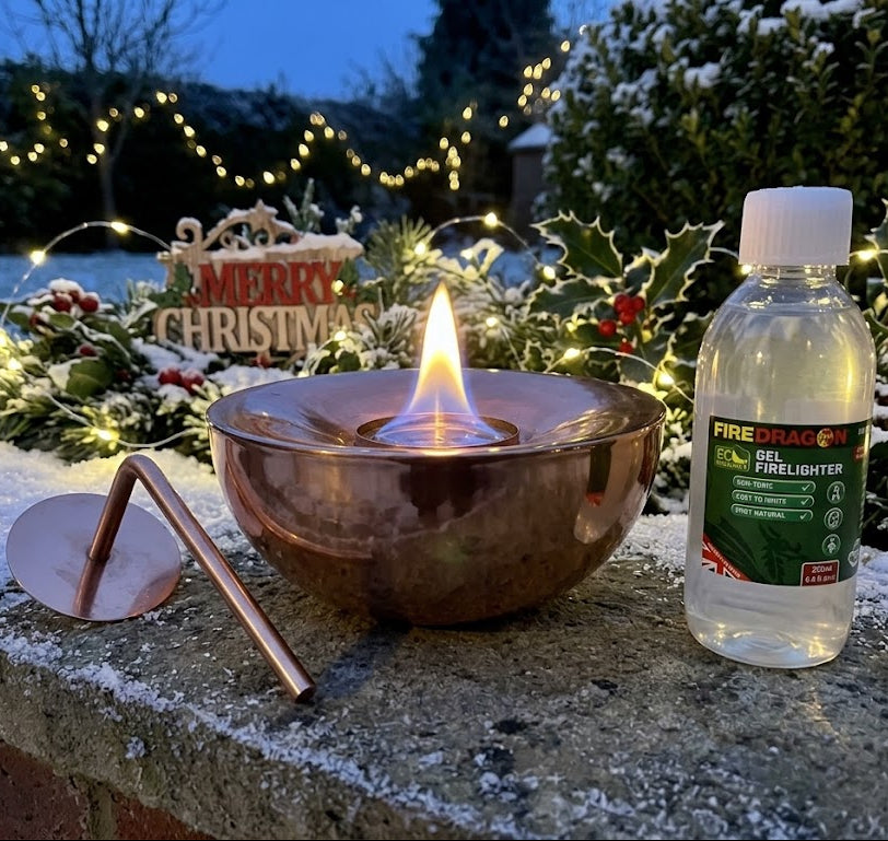 A copper-colored metal Gel Lamp with a lit flame sits on a snowy stone ledge outdoors at dusk, alongside a bottle of Fire Dragon Fire Starter fluid and a metal snuffer. Decorative holly, snow, and warm string lights are blurred in the background.