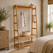 A tall wooden storage rack with ladder-style side supports and two slatted shelves, shown in a styled bedroom. The rack holds a white top, striped towel, hat, basket, and shoes, and is placed beside a bed and window.
