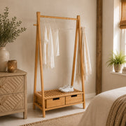 A light natural wood A-frame clothing rack with two drawers at the base, shown in a styled bedroom setting. Natural wooden sideboard and plants decorating the room.