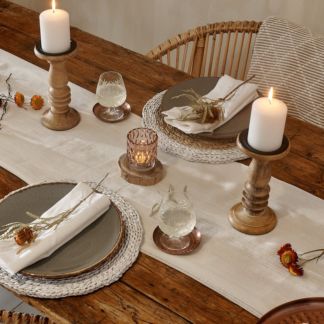 Wooden candle holders on cotton runner on wooden table, with white rattan place mats, plates and rattan chair