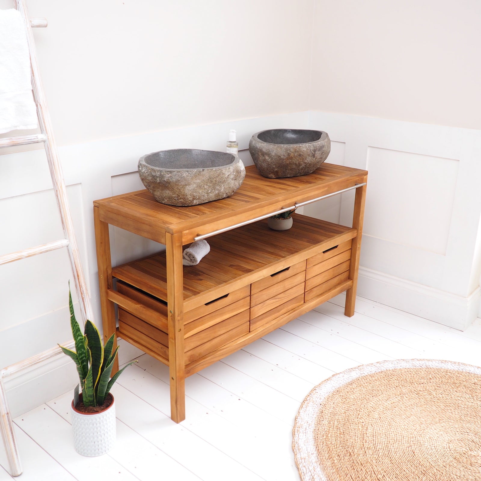 A teak wooden bathroom console table with two stone vessel sinks on top, styled in a bright white-walled bathroom corner with a plant, ladder, and woven rug.
