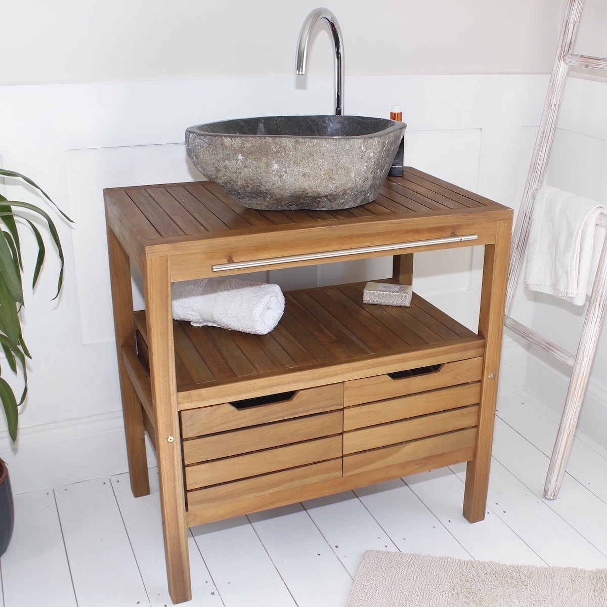 A teak wooden bathroom vanity cabinet with a slatted front and open shelf, shown with a stone vessel sink, curved faucet, rolled towel, and soap in a bright bathroom setting.