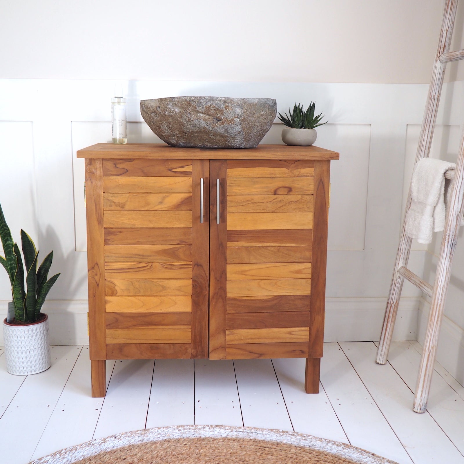 A wooden bathroom vanity cabinet with two doors and a matching wood top, styled with a stone vessel sink, small plants, and a white towel on a ladder in a bright room.