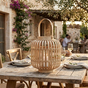 A natural bamboo lantern with a top handle sits on a rustic wooden dining table set for a meal outdoors in a sunny, elegant patio setting.