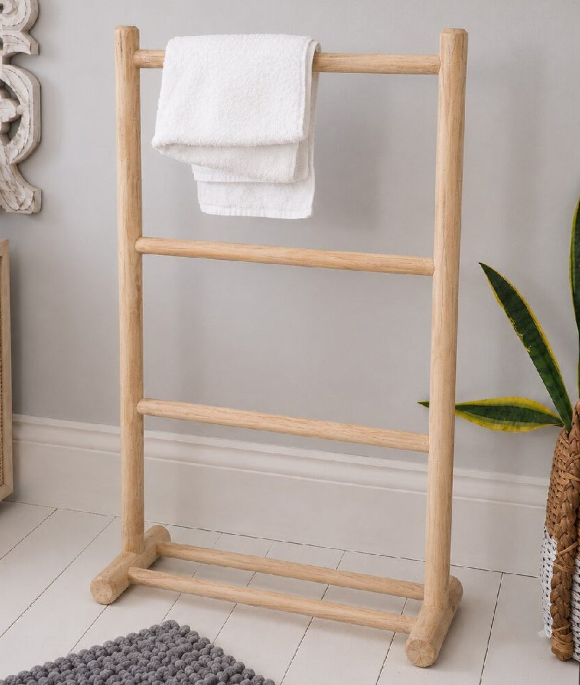 A natural light wood four-tier towel rack with a folded white towel, set in a styled bathroom corner with a gray wall, white tiled floor, and decorative plants.