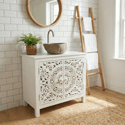 A white bathroom vanity cabinet with ornate carved doors, styled with a stone vessel sink, a curved faucet, a potted green plant, a round wood-framed mirror, and a wooden towel ladder in a bright bathroom setting.