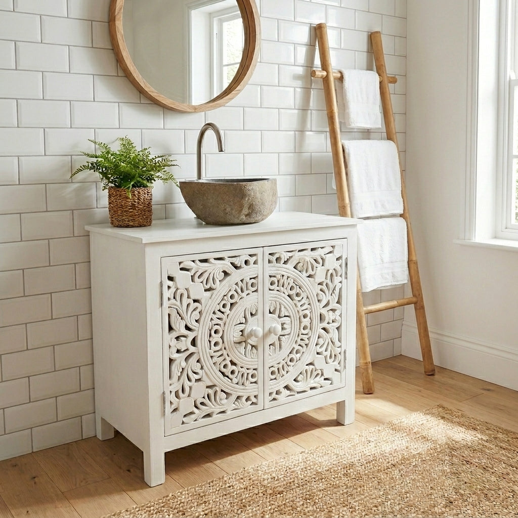 A white bathroom vanity cabinet with ornate carved doors, styled with a stone vessel sink, a curved faucet, a potted green plant, a round wood-framed mirror, and a wooden towel ladder in a bright bathroom setting.