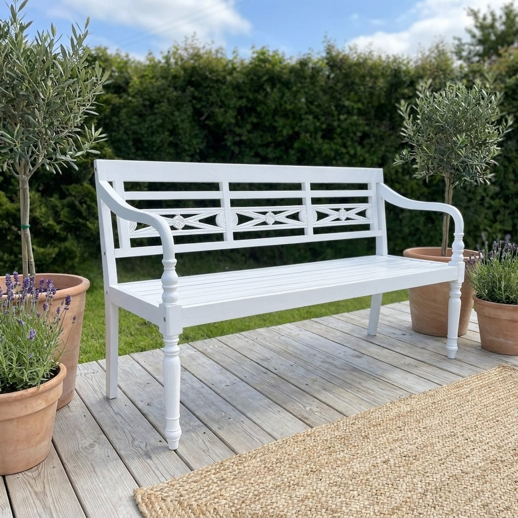 A white wooden garden bench with turned legs and decorative slat backrest, placed on a weathered wooden deck outdoors with potted plants and green hedges in the background.