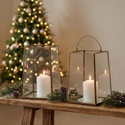 Two glass lantern candle holders with metal frames sit on a rustic wooden table, each holding a lit white pillar candle. Pine branches, pinecones, and a decorated Christmas tree appear in the background against a warm beige wall.
