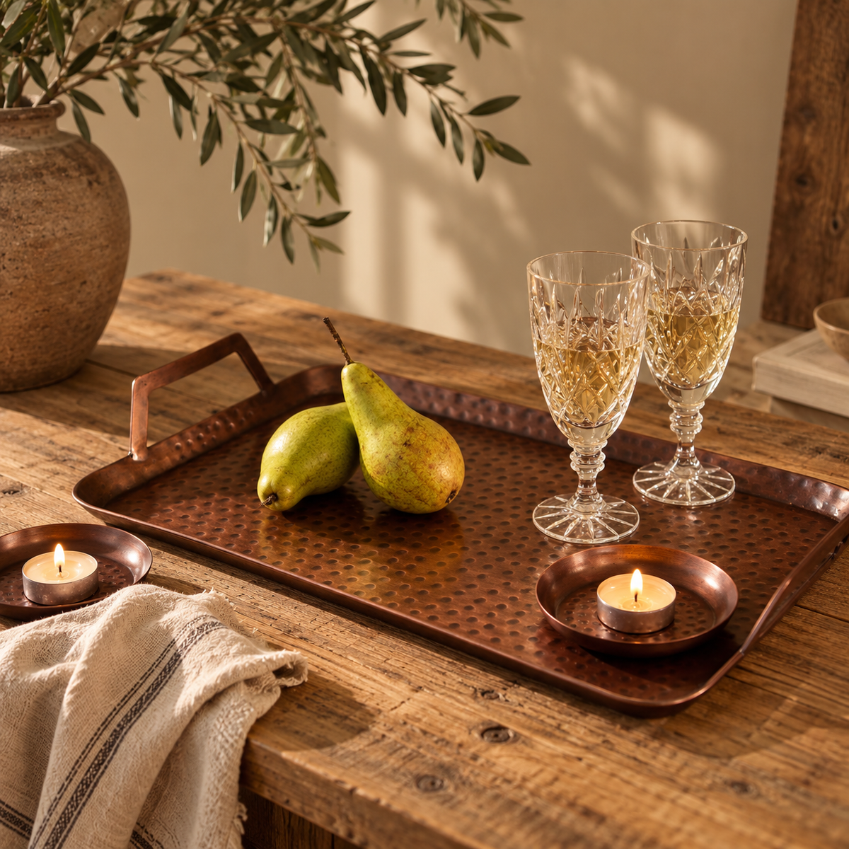 A copper serving tray with handles is styled on a rustic wooden table with matching antique copper coasters, holding two pears, two clear cut-glass wine glasses, and two small lit tea lights. 