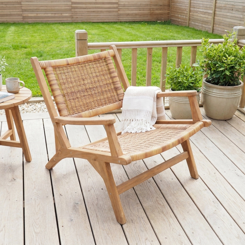 A light natural wood outdoor lounge chair with woven cane back and seat, shown on a wooden deck with a white throw blanket draped over the arm. A small matching side table is partially visible on the left, with a green lawn and fence in the background.
