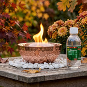 A hammered copper fire bowl with a visible flame sits on white stones beside a bottle of Fire Starter fuel lighter on a wooden table, with autumn foliage in the background.