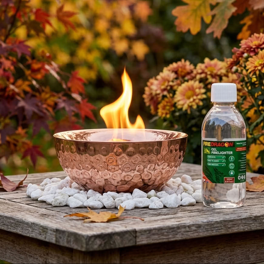 A hammered copper fire bowl with a visible flame sits on white stones beside a bottle of Fire Starter fuel lighter on a wooden table, with autumn foliage in the background.