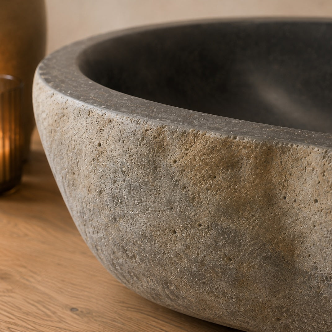 A close-up of a round stone vessel sink with a textured, porous surface in warm gray and beige tones, set on a wooden surface.