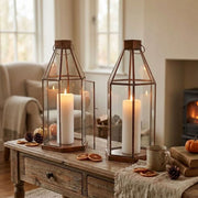Two brass metal candle lanterns, placed on a wooden table in a cozy living room setting with fall decor.