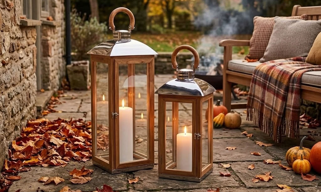 Two wooden lanterns on a garden patio, lit with white pillar candles, autumn leaves, pumpkins, a bench, and a plaid throw blanket in the background.