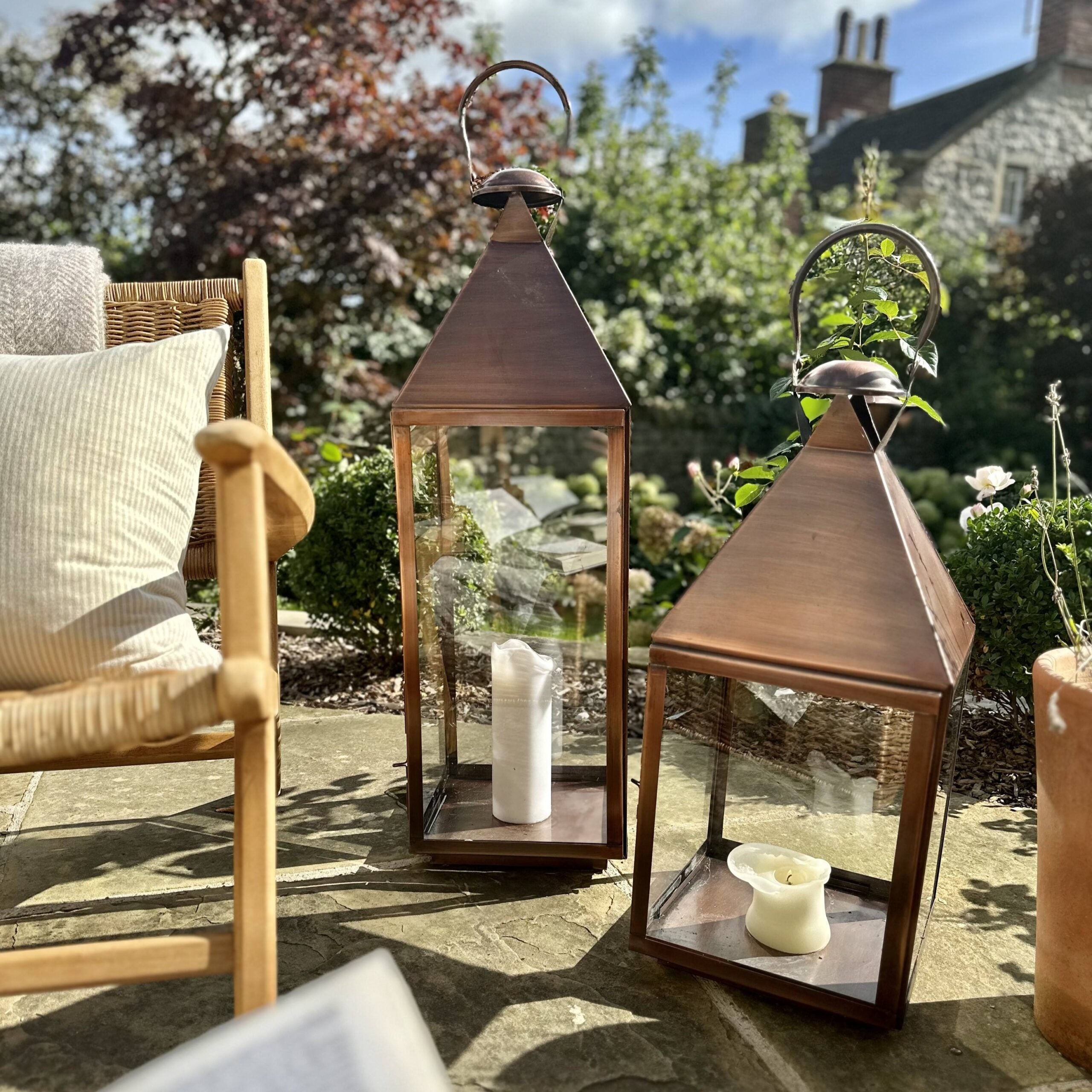 Two aged copper lanterns outdoors in garden setting, next to rattan chair on sunny patio