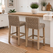 Two woven wicker bar stools with light wood legs, positioned at a kitchen island with a wooden countertop in a bright, modern kitchen.