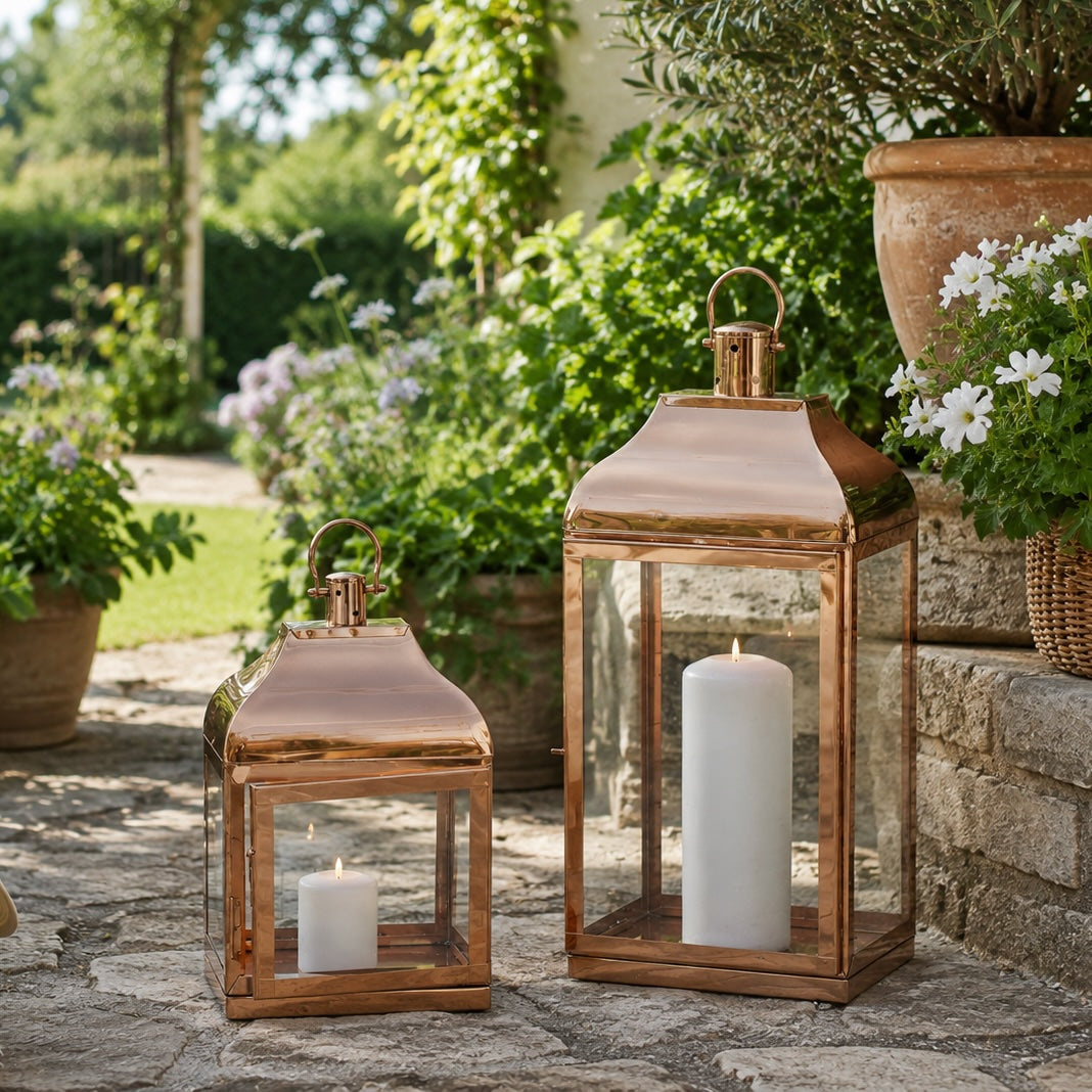 Two rose gold lanterns in a garden setting surounded by plants with a lawn in the background
