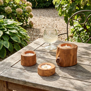 Three small wooden candle holders shaped like tree stumps are arranged on a weathered wooden table outdoors, with a glass and garden foliage in the background.