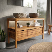 A wooden bathroom vanity console with open shelving and multiple drawers, styled in a modern bathroom setting with stone vessel sinks, towels, and plants.
