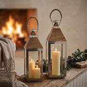 Two decorative lanterns with rope handles and wood tops, displayed on a rustic table in a cosy room with a fireplace in the background.