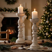 Two distressed white wooden candlestick holders, styled on a rustic table with festive accessories. In front of a cozy fireplace and Christmas tree background.