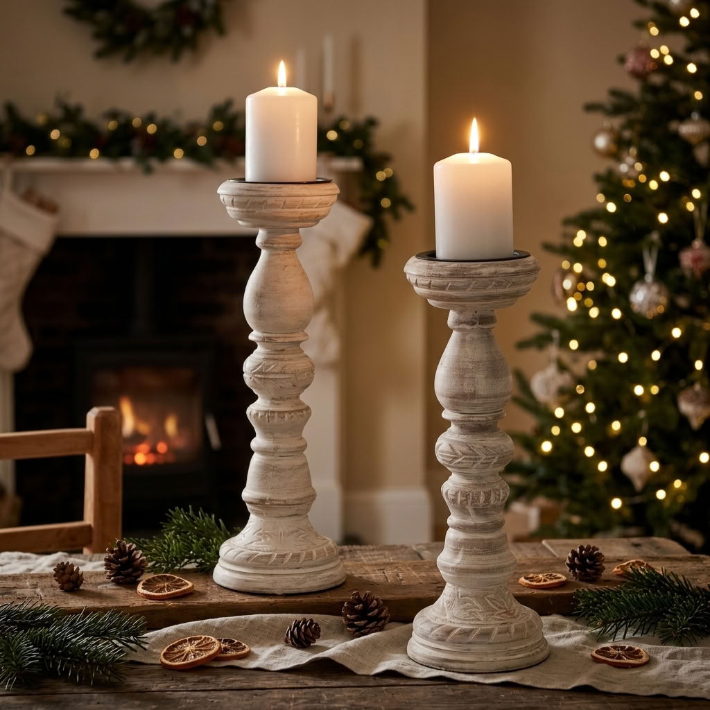 Two distressed white wooden candlestick holders, styled on a rustic table with festive accessories. In front of a cozy fireplace and Christmas tree background.
