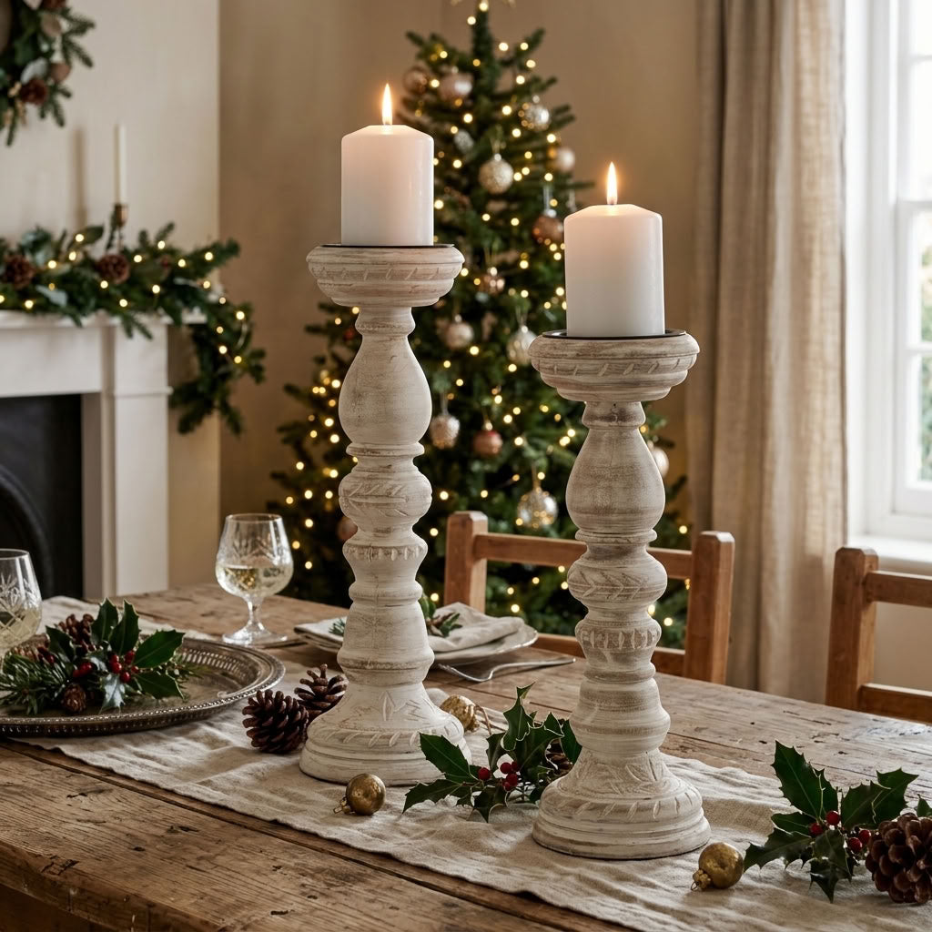Two tall white candlestick holders with lit on a reclaimed wooden dining table, decorated with holly, pinecones, and a Christmas tree in the background.