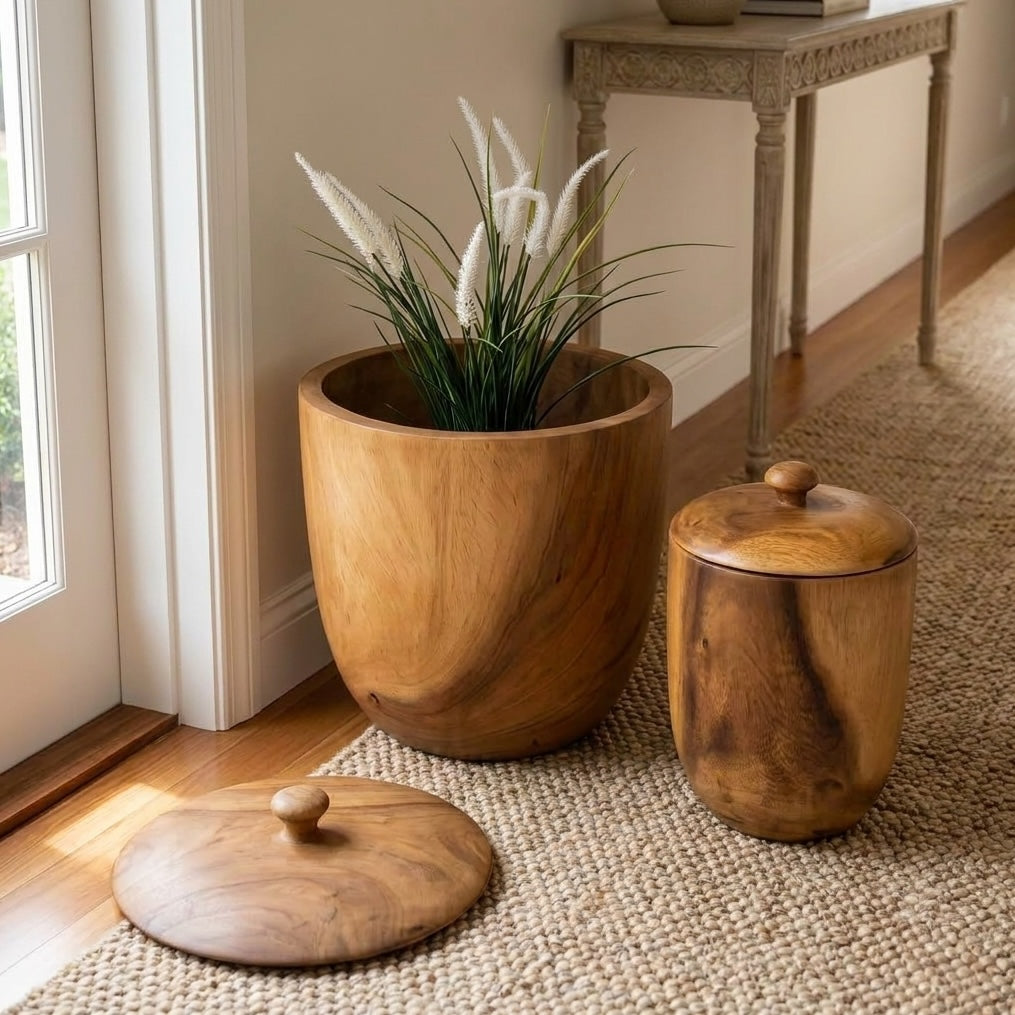 Two round wooden pots with lids, one large and one small, styled on a woven rug next to a window in a home interior. The larger wood pot is shown holding a green plant.